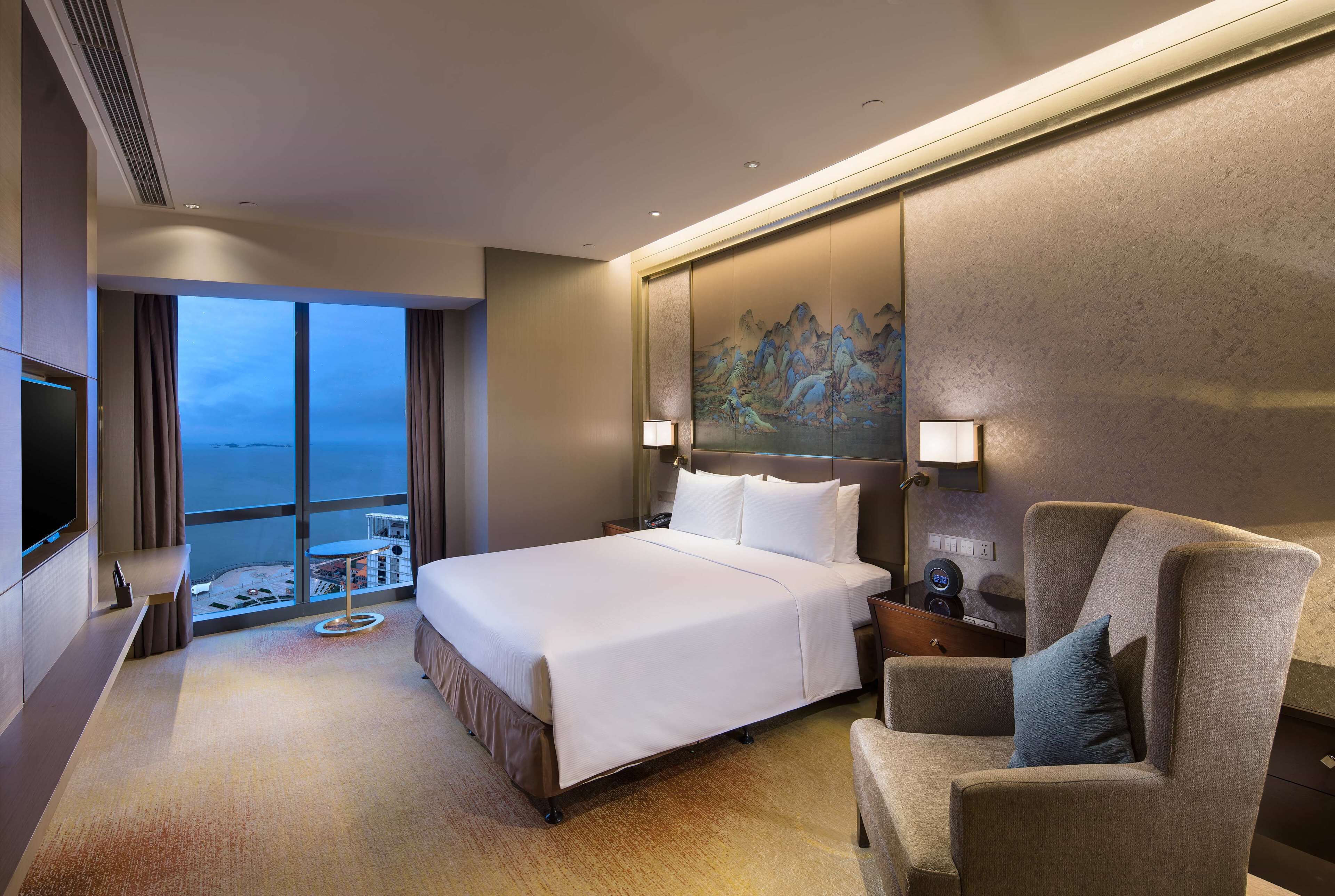 A hotel room with a large bed, a seating area, a TV, and a view of the ocean.