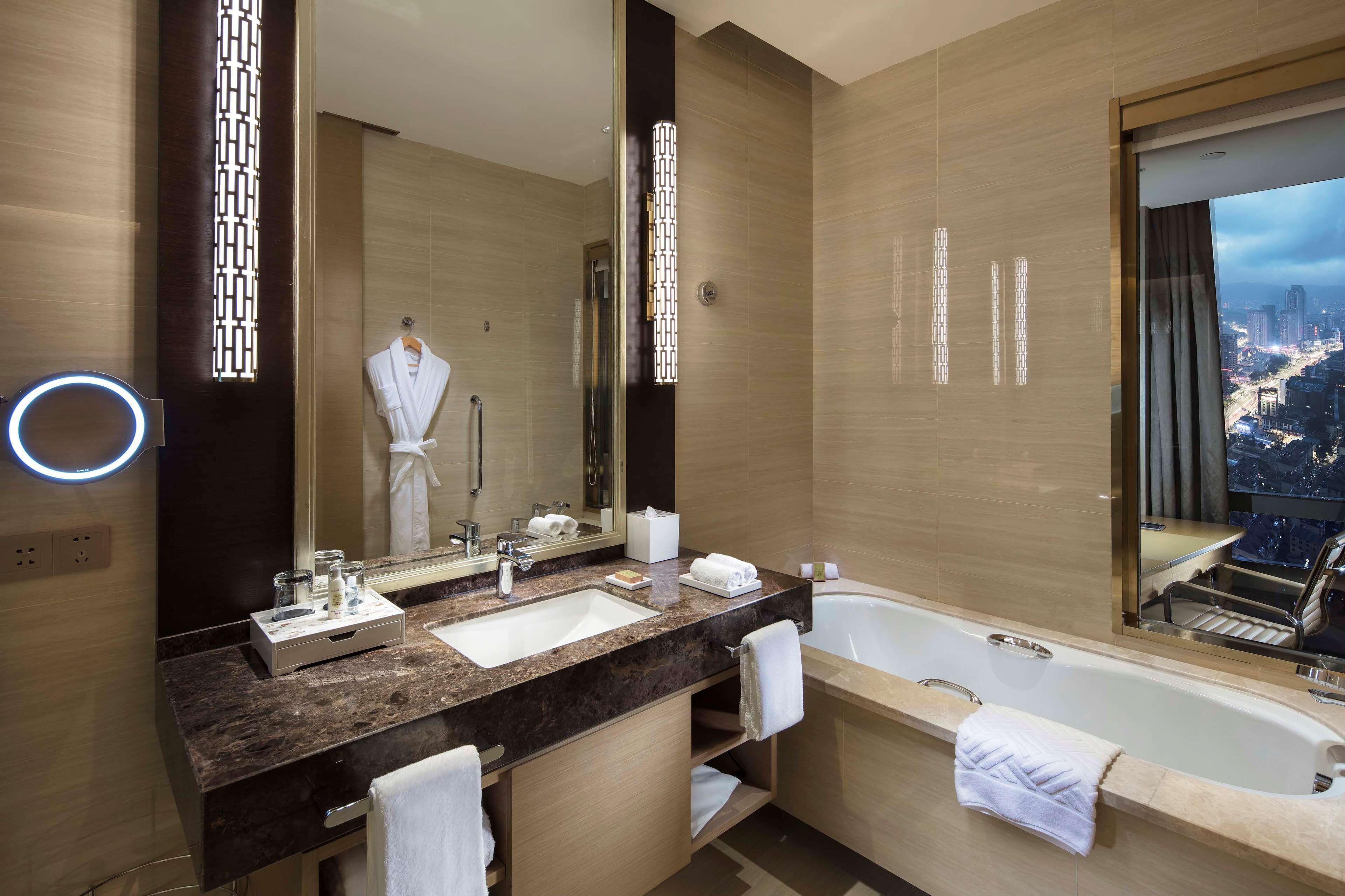 A modern bathroom with a large mirror, a sink, a bathtub, and a view of the city through a large window.