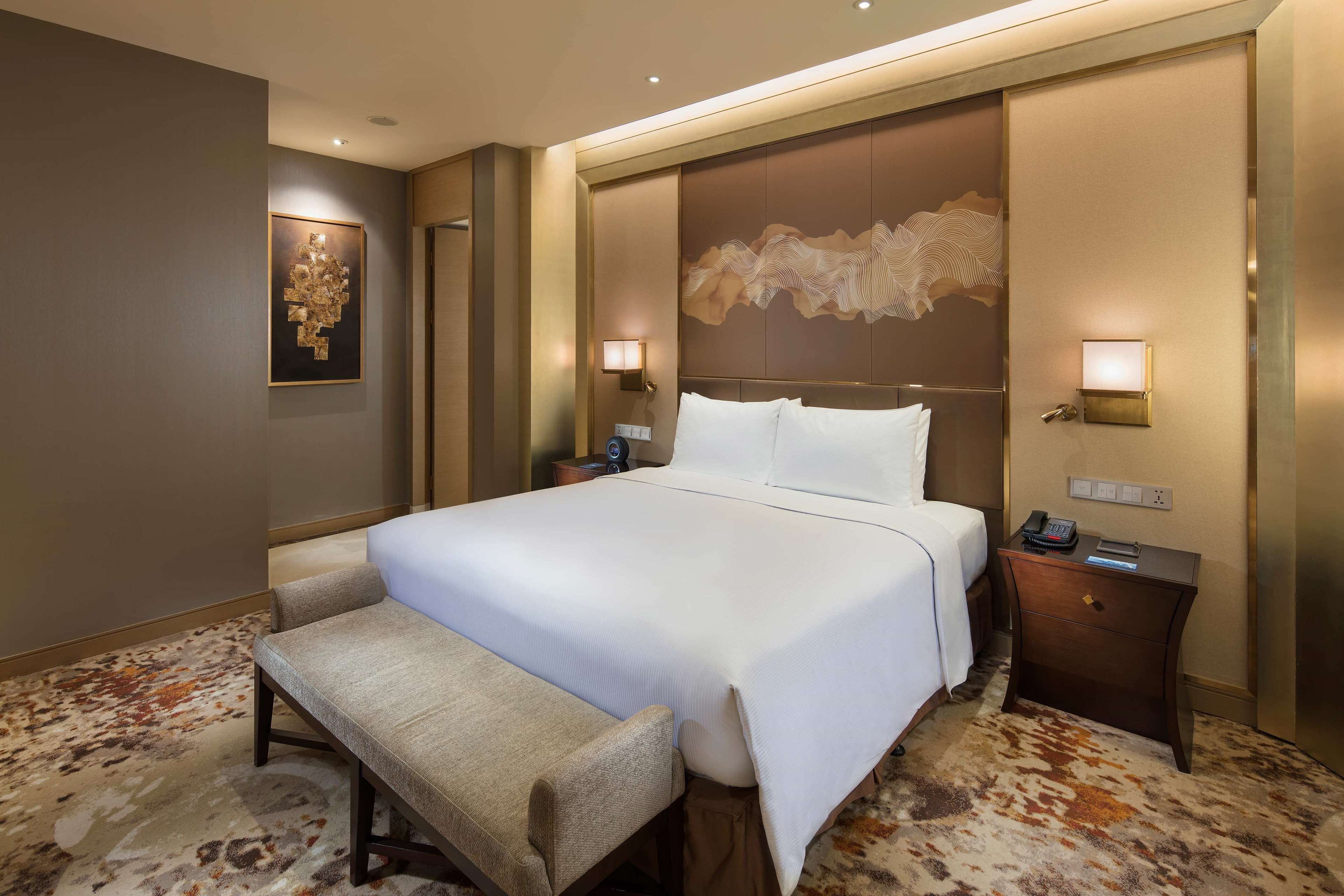 A hotel room with a large bed, a bedside table with a lamp, a chair, and a wall-mounted artwork.