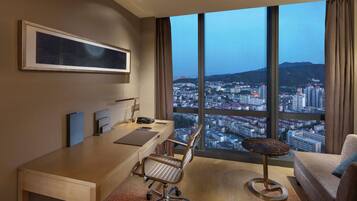 Habitación de hotel moderna con un ventanal amplio que ofrece vistas a la ciudad, un escritorio con silla, un sofá y una mesita redonda.