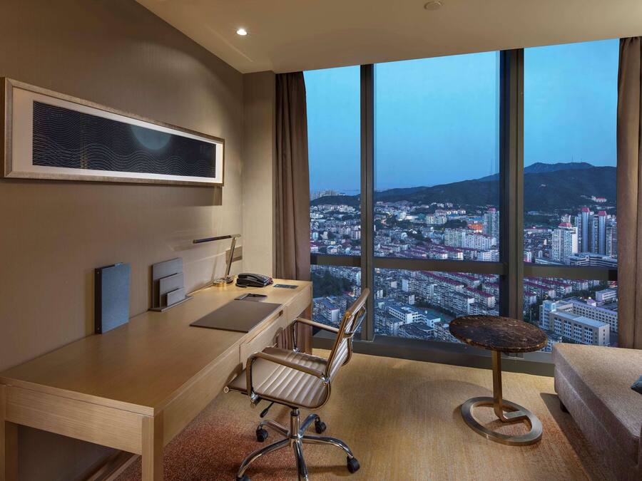 Habitación de hotel moderna con un ventanal amplio que ofrece vistas a la ciudad, un escritorio con silla, un sofá y una mesita redonda.