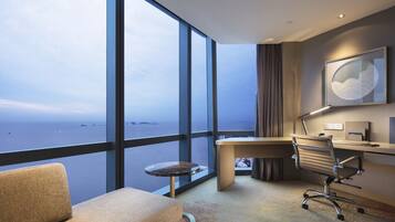 A modern hotel room with a large window offering a view of the sea, a desk with a chair, and a small round table.
