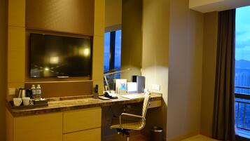 A hotel room with a desk, chair, TV, and a view of the cityscape through the window.