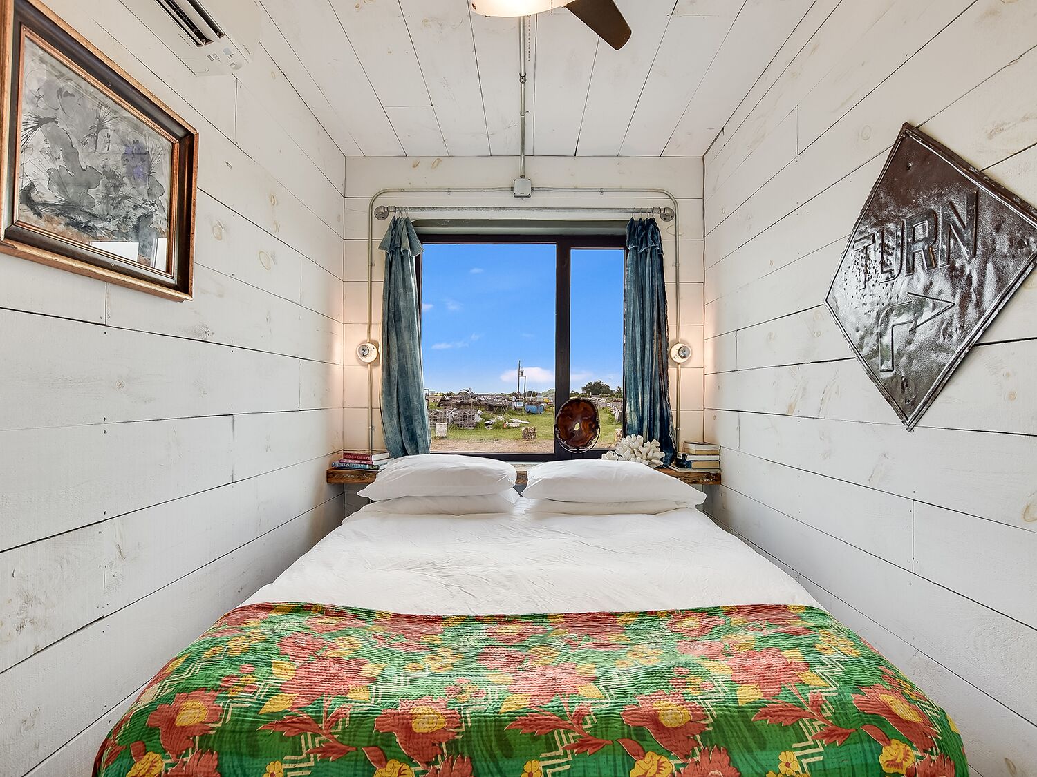 A bedroom with a large bed, a window with curtains, a ceiling fan, and a framed picture on the wall.