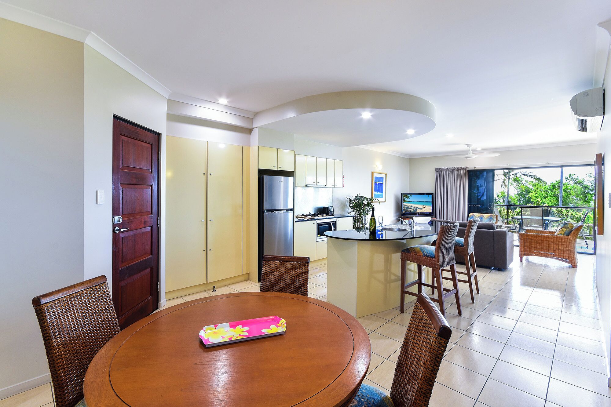 A modern kitchen with a central island, dining area with wicker chairs, and a balcony with a view.