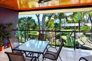 Un balcón con una mesa y sillas de vidrio, con vistas a un jardín exuberante y al mar.