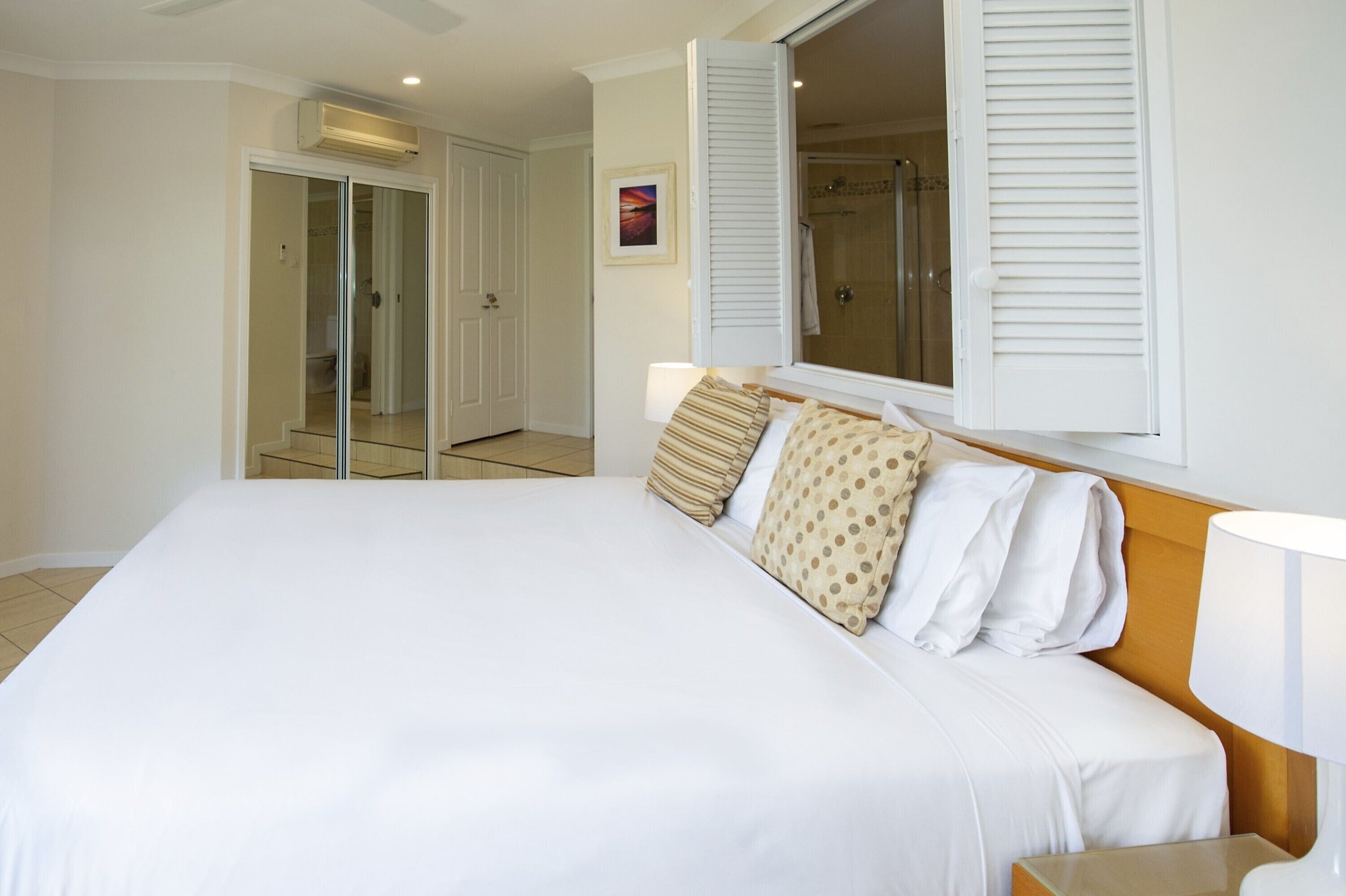 A hotel room with a large bed, a wooden headboard, a window with shutters, a mirror, and a door leading to a bathroom.