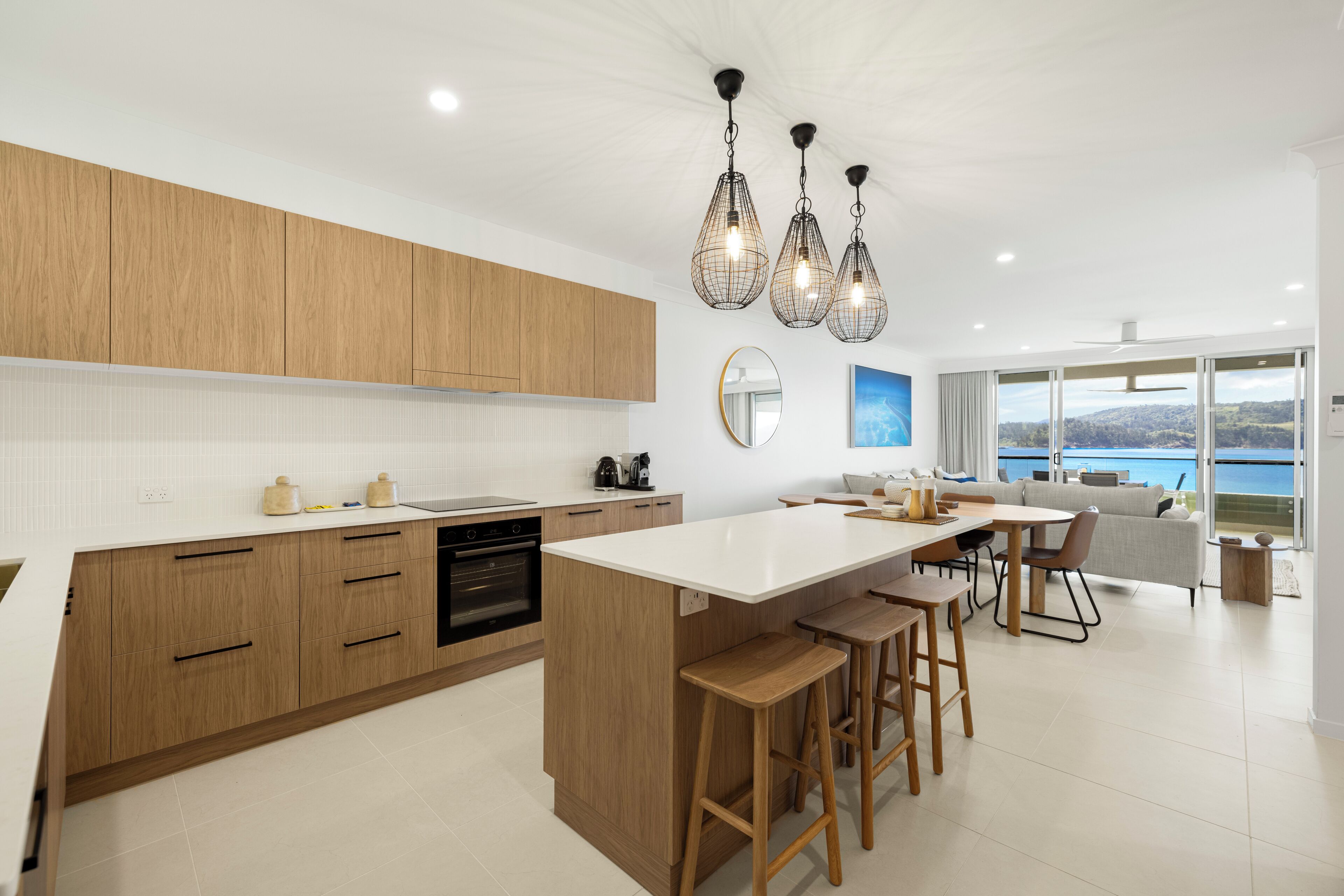 A modern kitchen with wooden cabinets, a central island with stools, pendant lighting, and a view of the sea through large windows.
