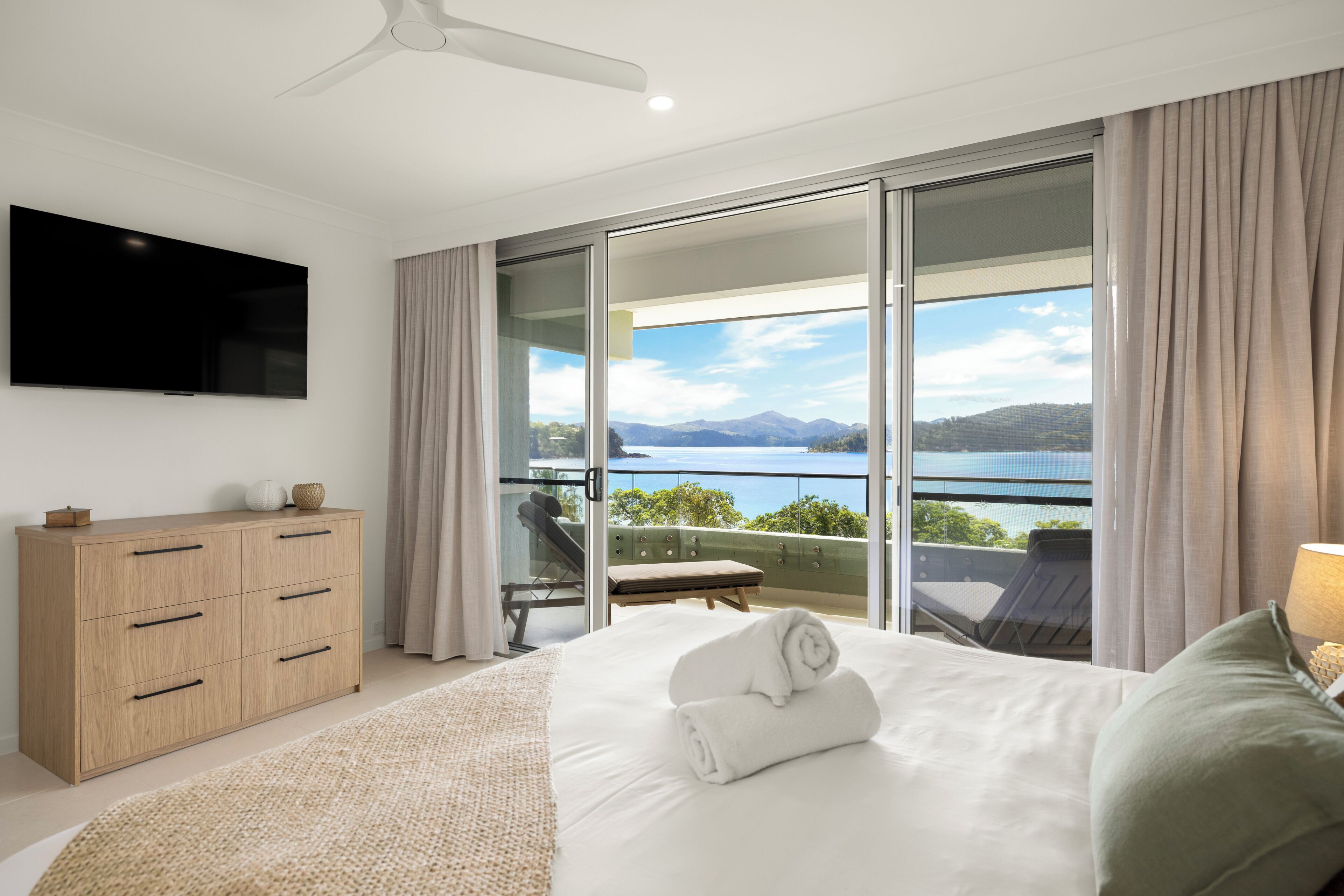 Ein Schlafzimmer mit Bett, Fernseher, einem Holzschrank und einem Balkon mit Blick auf das Wasser.