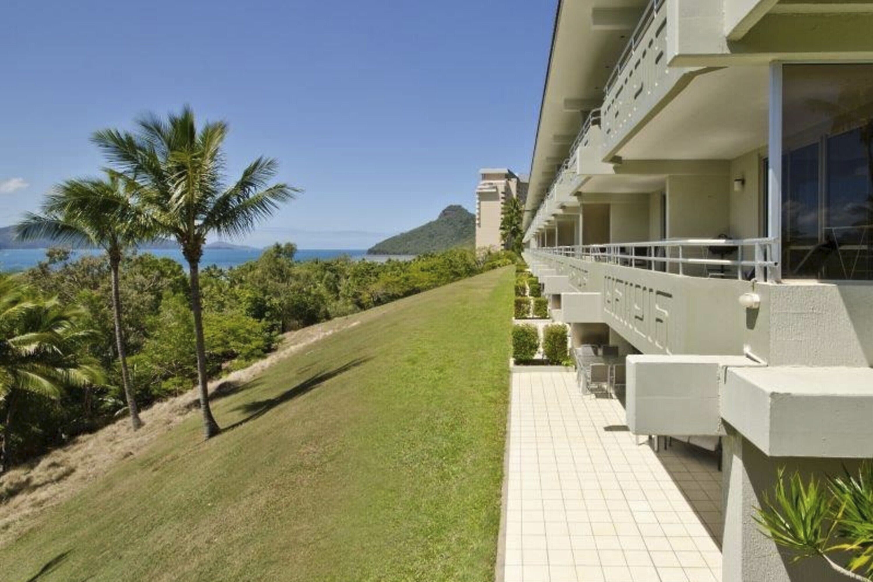 A hotel with a balcony overlooking a grassy area with palm trees and a view of the ocean.