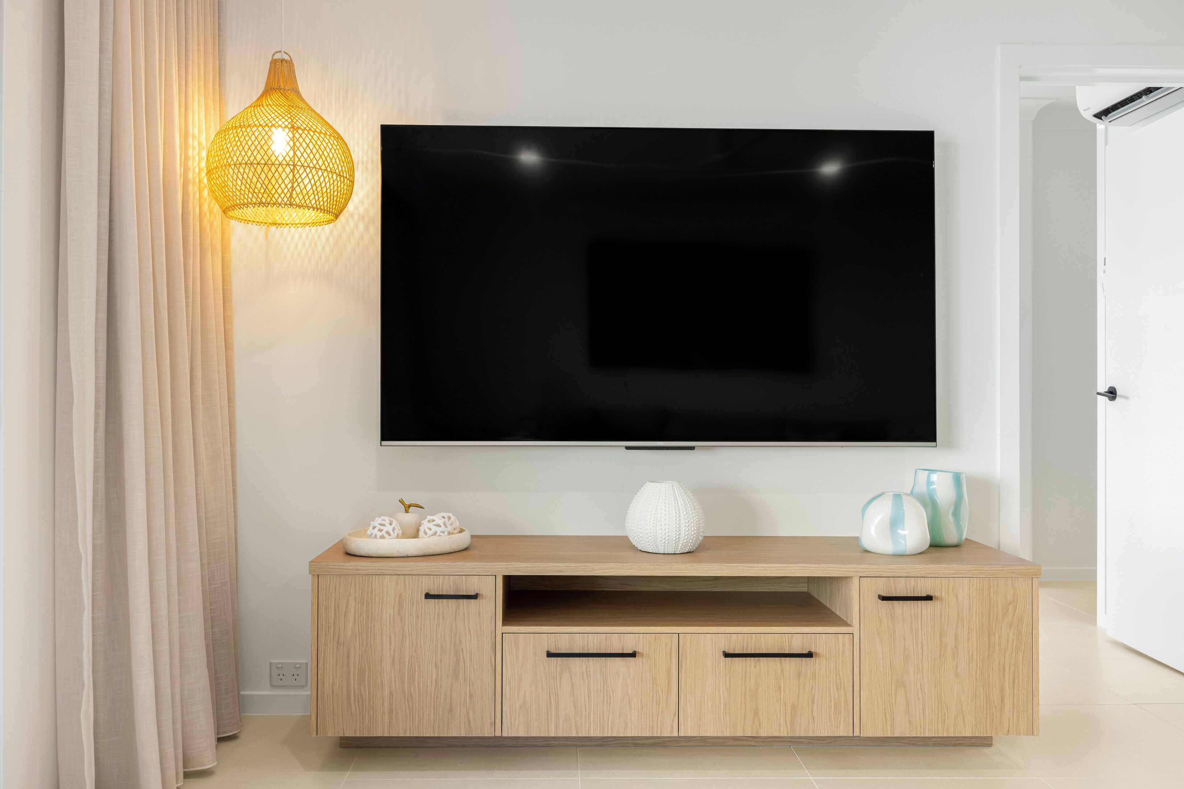Un saló modern amb un televisor de pantalla plana muntat a la paret, un moble de roba baix amb calaixos de fusta i una llum penjant de fibra natural.