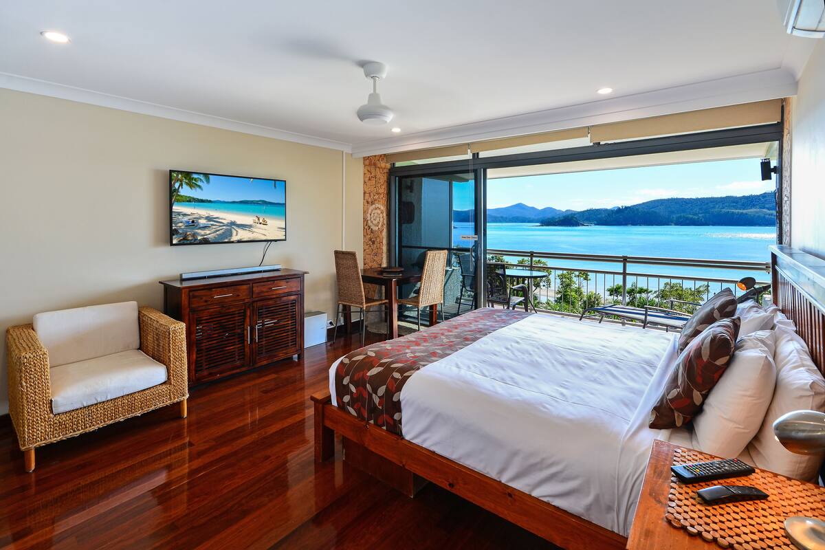 Une chambre dotée d’un grand lit, d’un canapé, d’une télévision et d’un balcon offrant une vue sur un lac et des montagnes.