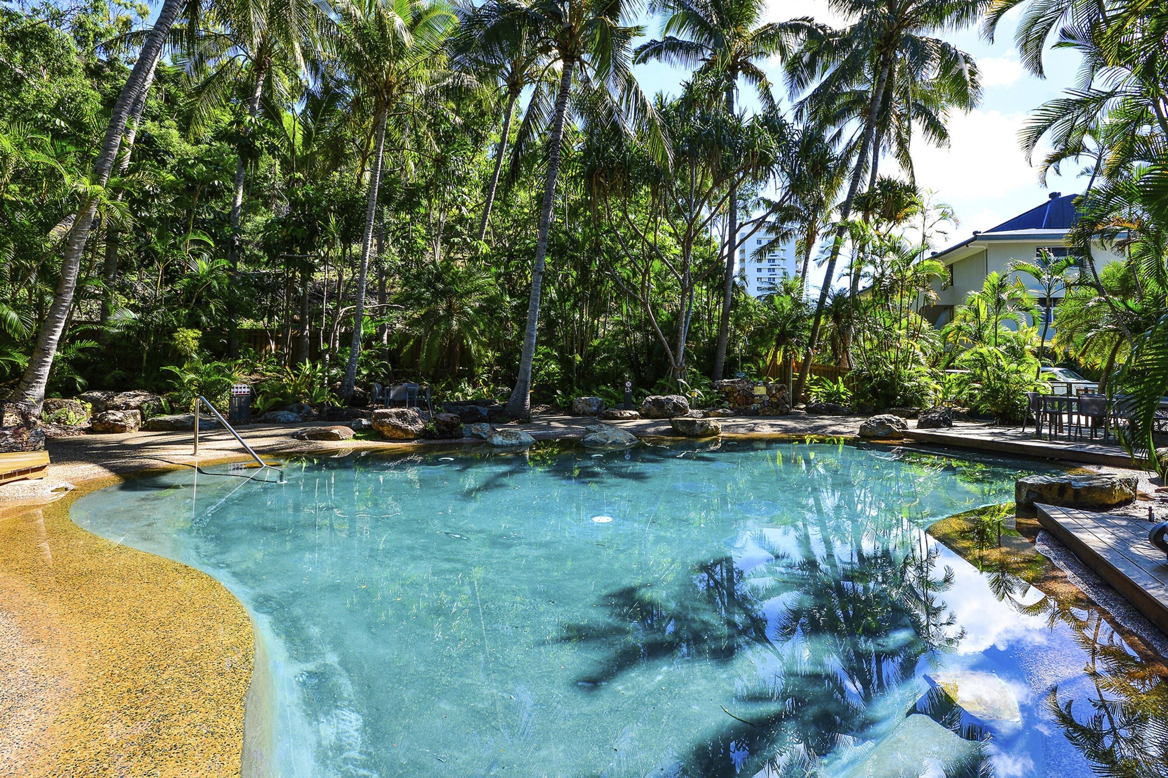 Una piscina cristalina rodeada de palmeras y rocas, con un edificio al fondo.