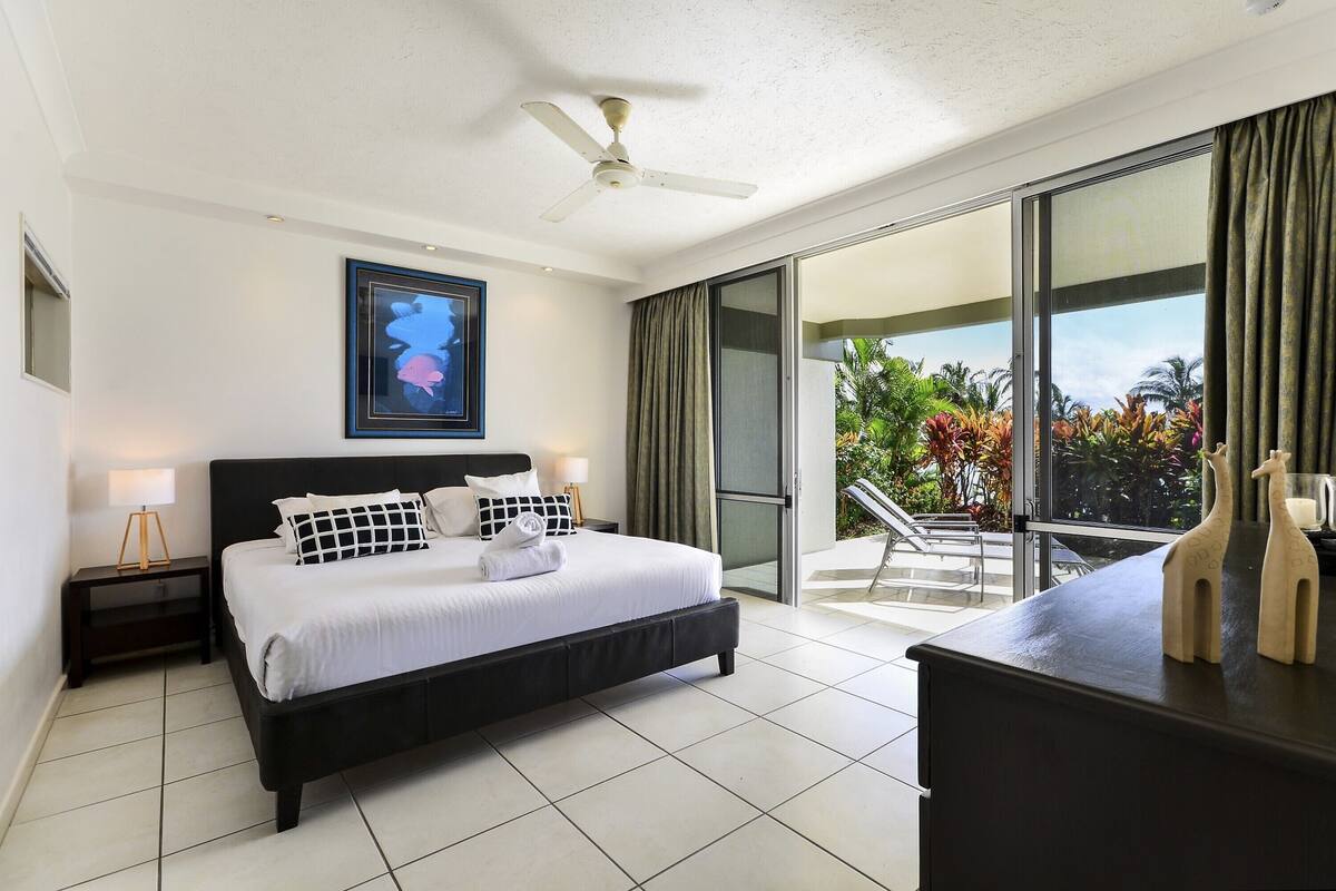 Une chambre à coucher comprenant un lit, une table de chevet, un ventilateur de plafond et donnant sur un balcon agrémenté de plantes.