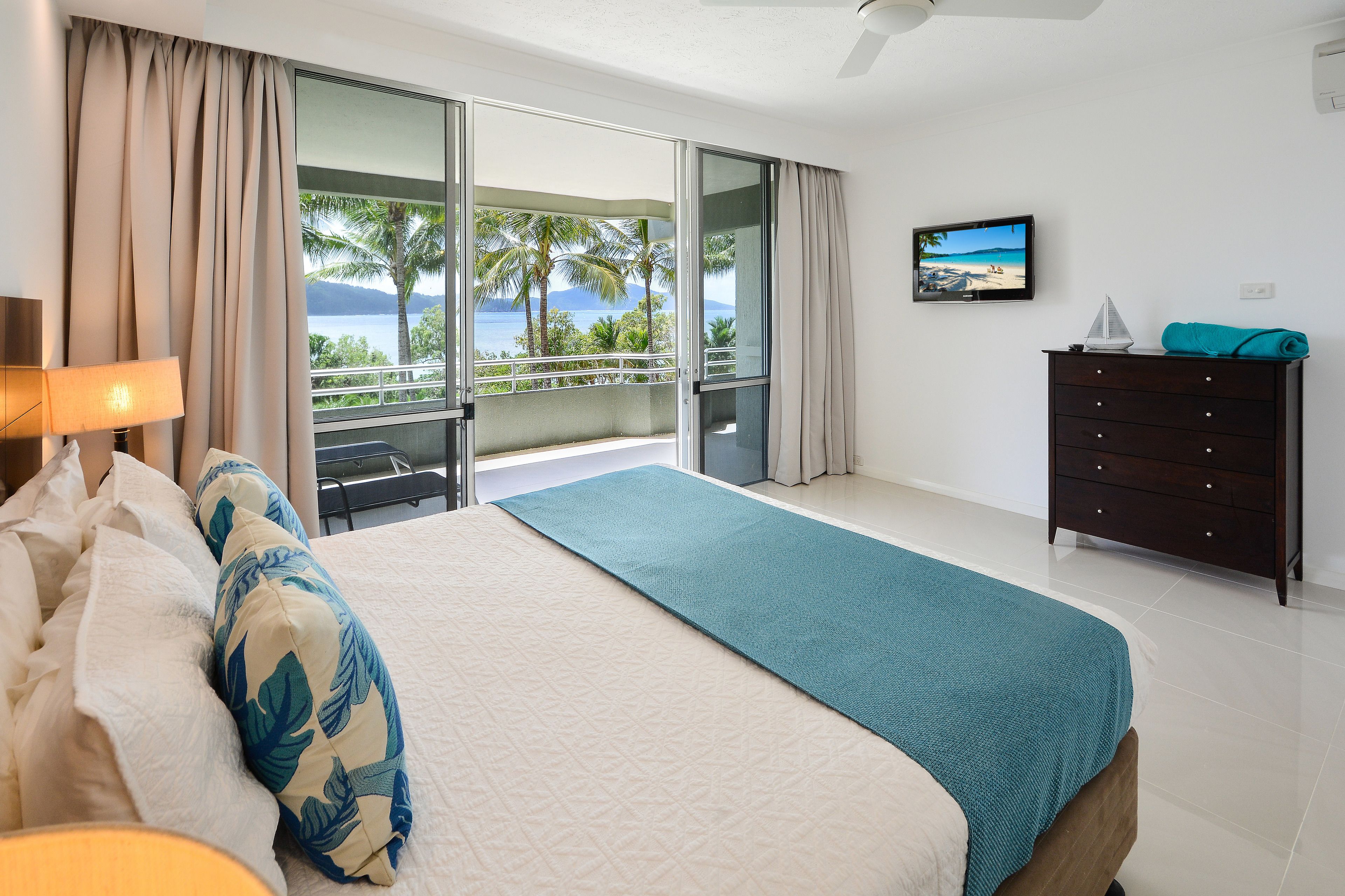 Hotelkamer met een bed, bureau, dressoir, televisie en balkon met uitzicht op zee en palmbomen.