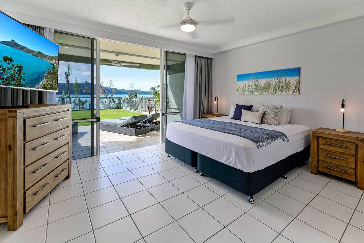 Une chambre avec un lit, une commode en bois, une télévision et un balcon donnant sur la mer.