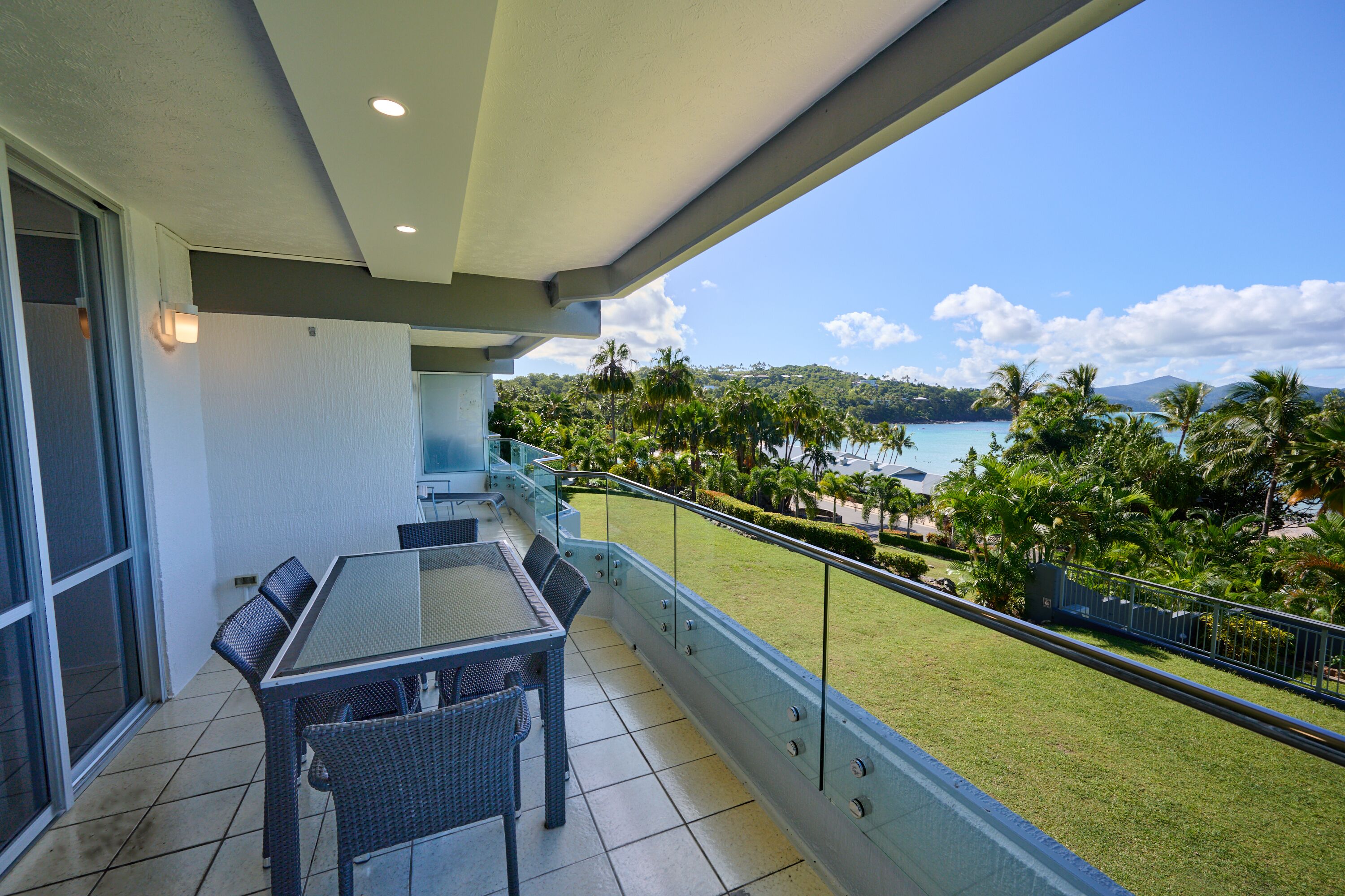 A balcony with a table and chairs, overlooking a grassy area with palm trees and a view of the water.