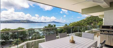 A balcony with a table and chairs, a grill, and a view of a lake and mountains.