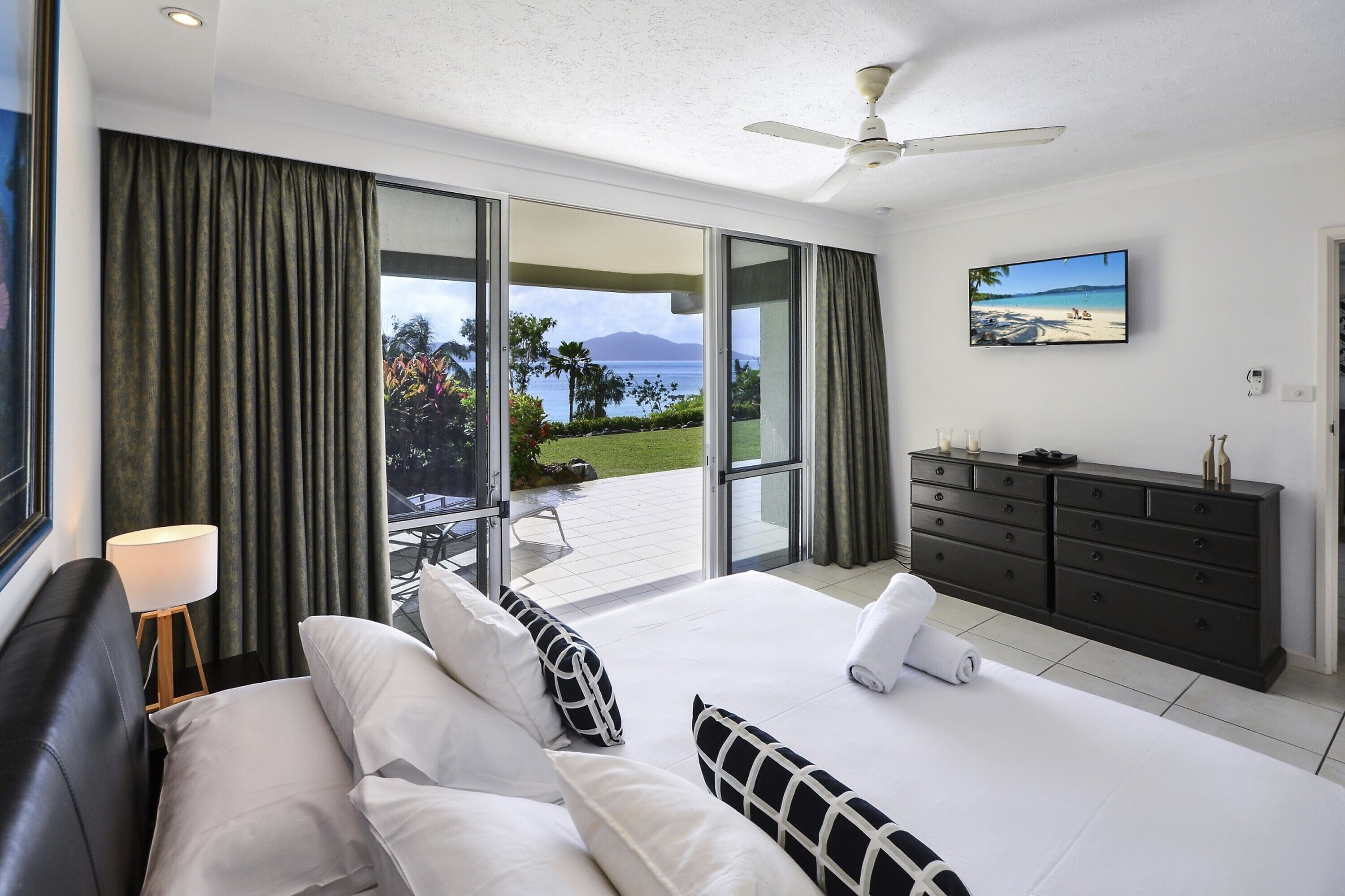 A modern hotel room with a large bed, a flat-screen TV, a dark wooden dresser, and a sliding glass door leading to a balcony with a view of the ocean and mountains.