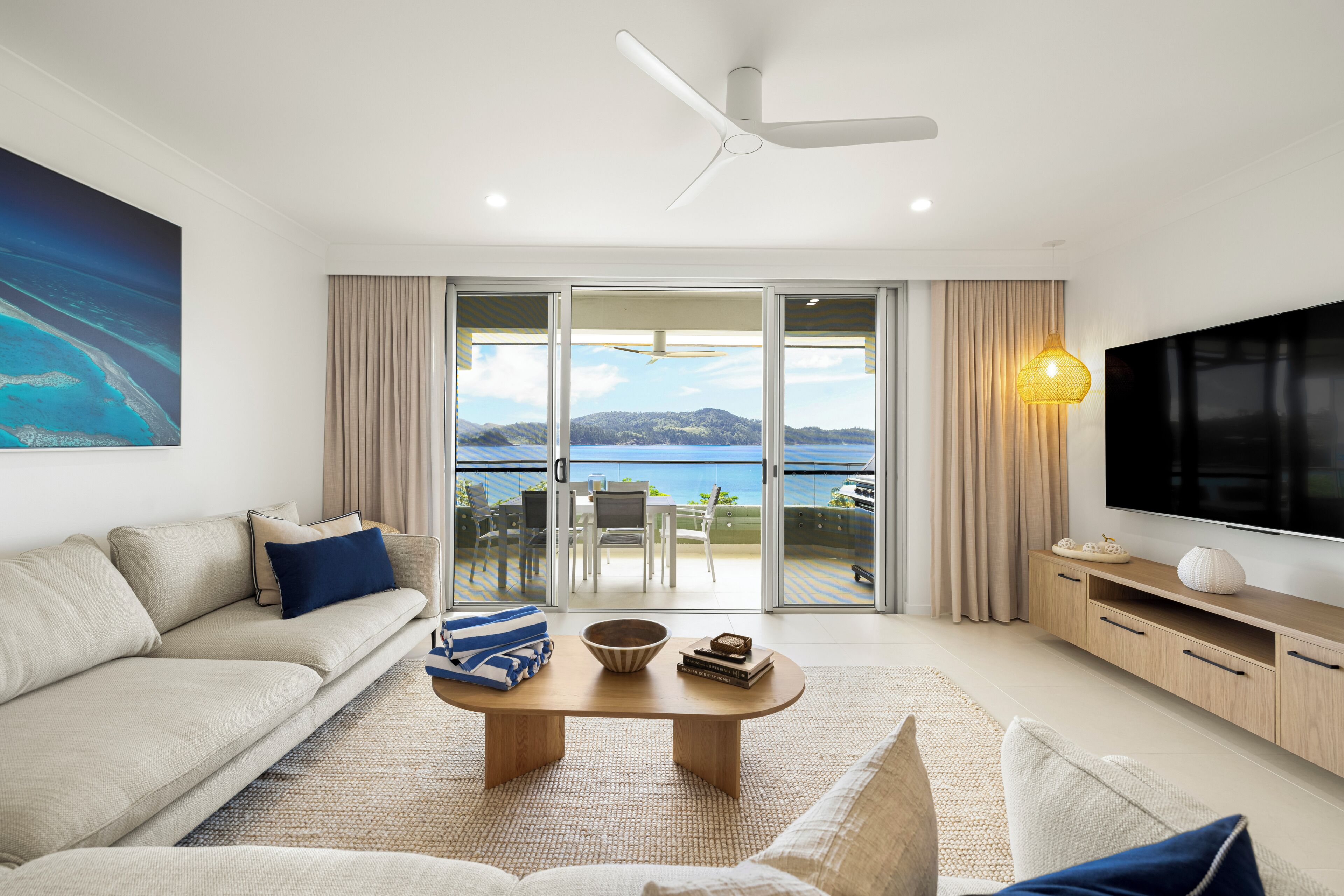 Ein modernes Wohnzimmer mit Sofa, Couchtisch und Fernseher, ein Balkon mit Meerblick und einem Deckenventilator.
