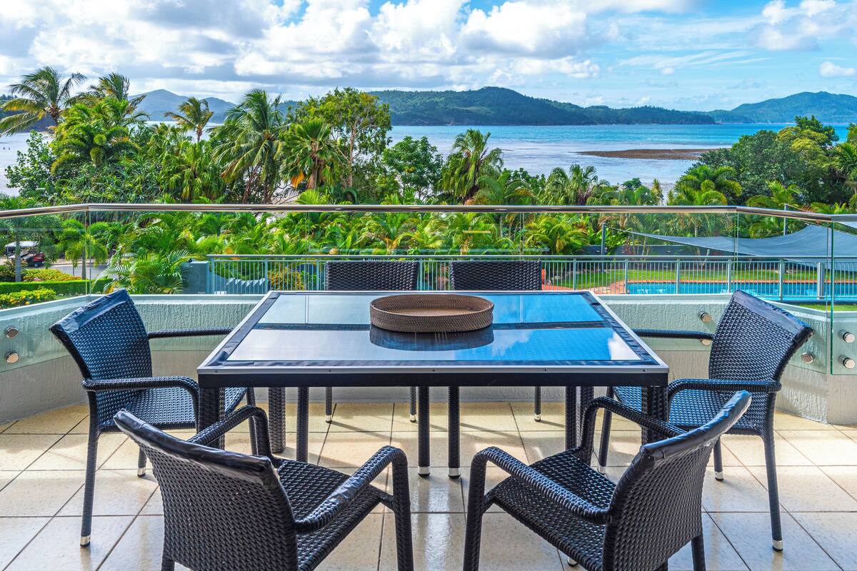 Un balcon avec une table et des chaises, donnant sur une plage et des montagnes.