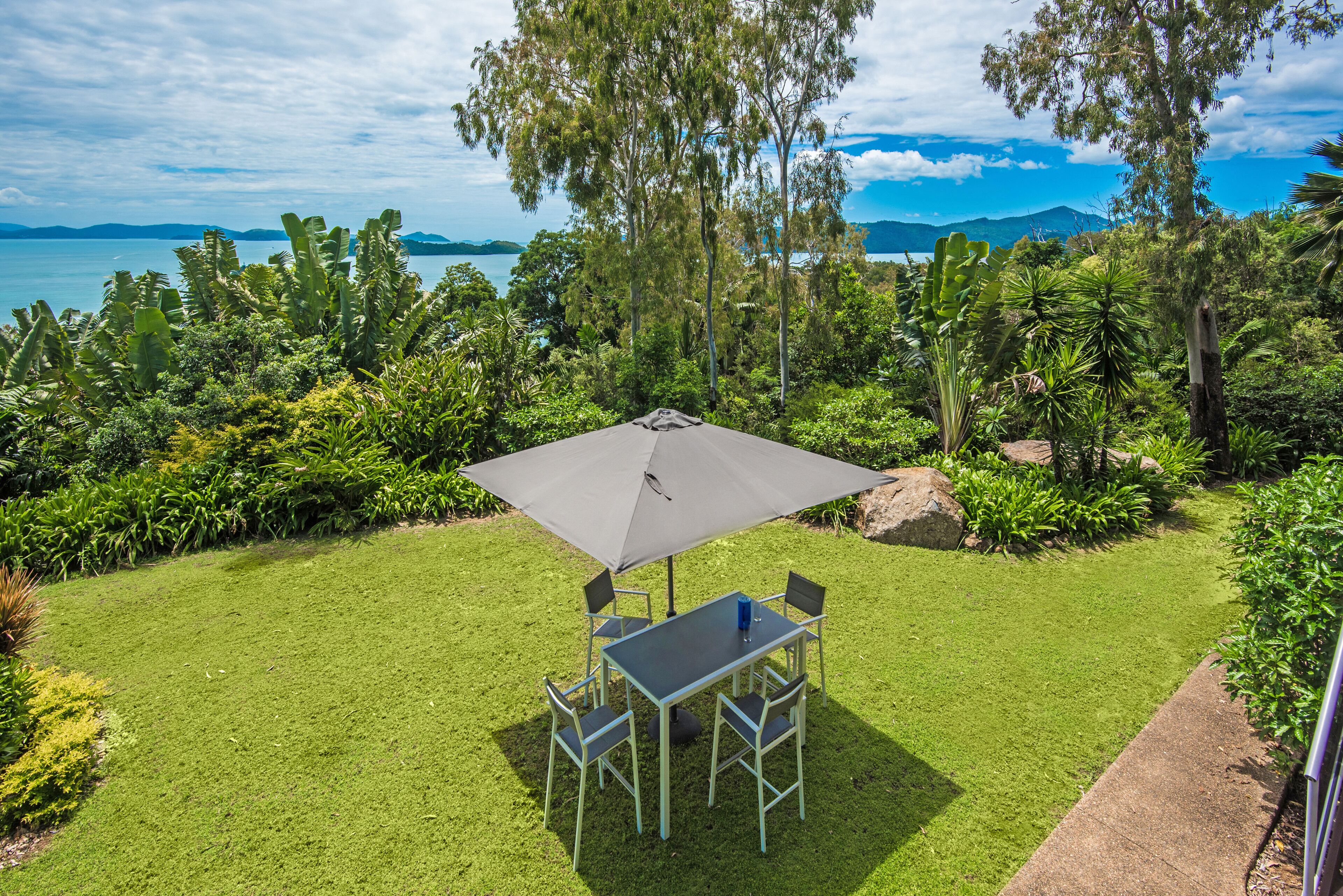 A patio with a table and chairs, surrounded by lush greenery and overlooking a body of water with mountains in the distance.