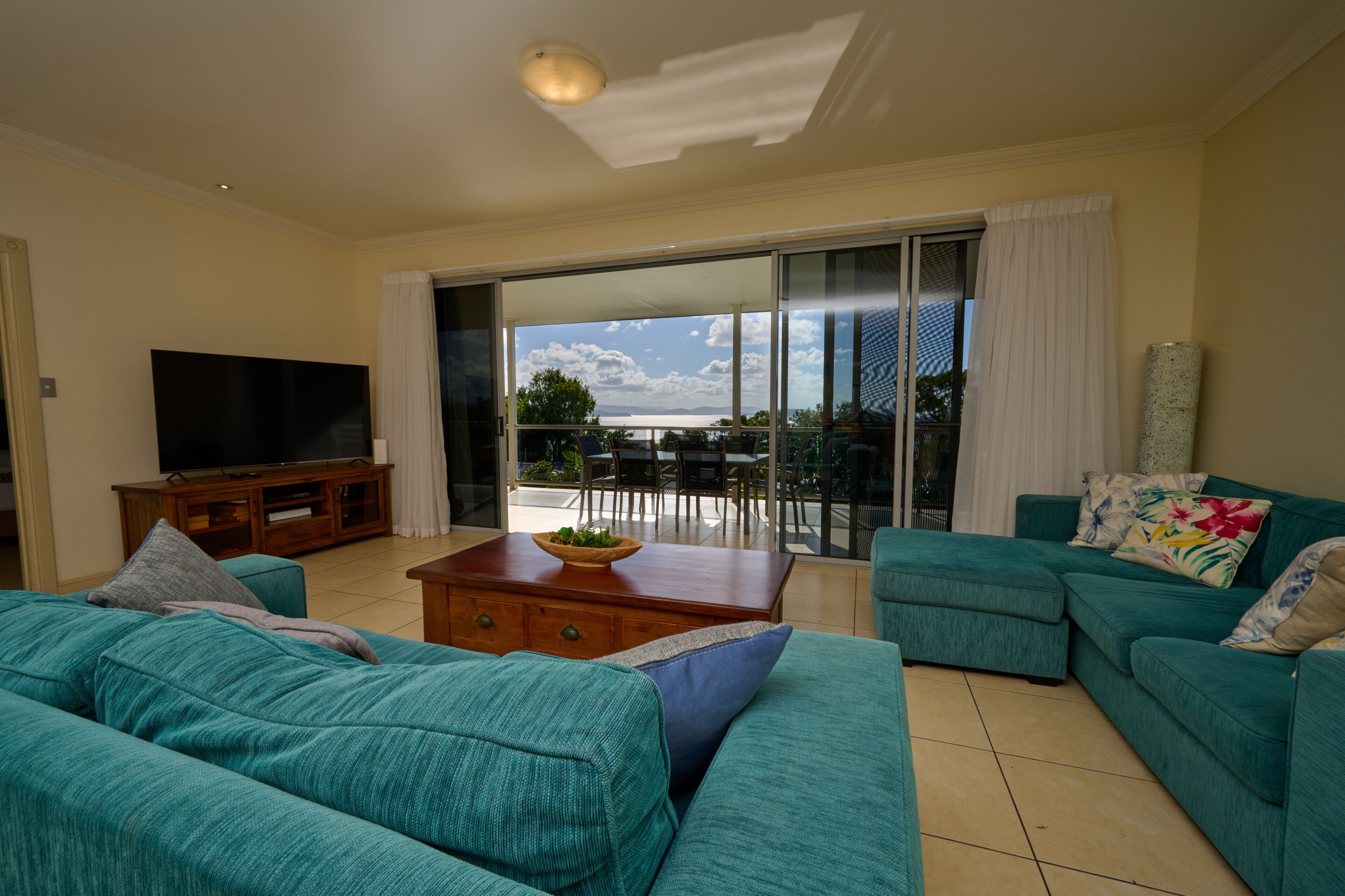A living room with teal sofas, a flat-screen TV, a wooden coffee table, and a view through a glass door to an outdoor patio.