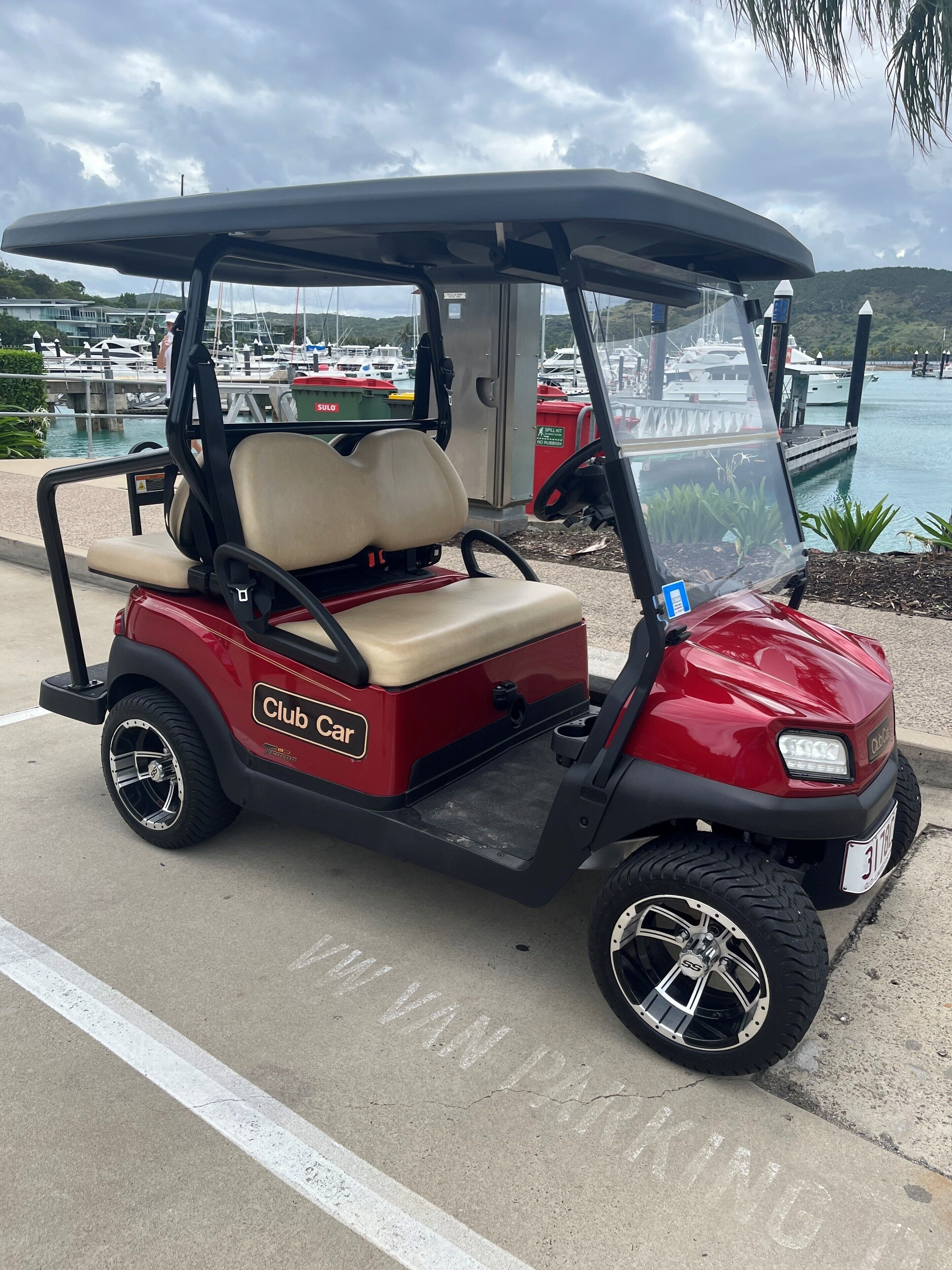 A red Club Car golf cart parked on a concrete surface with a marina and boats in the background.