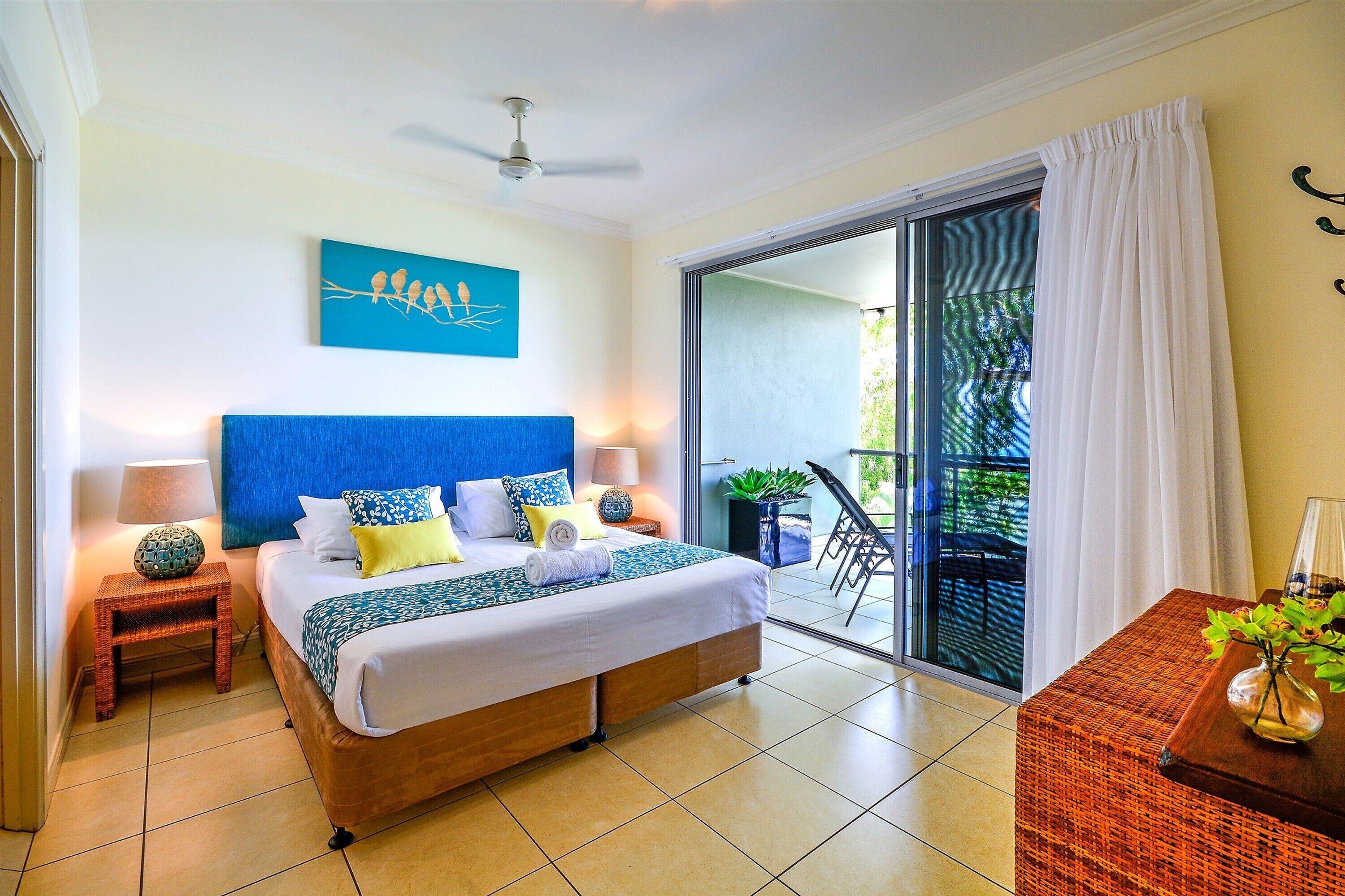 A well-lit bedroom with a large bed, a ceiling fan, a view of the outdoors through a sliding glass door, and a small table with a vase.