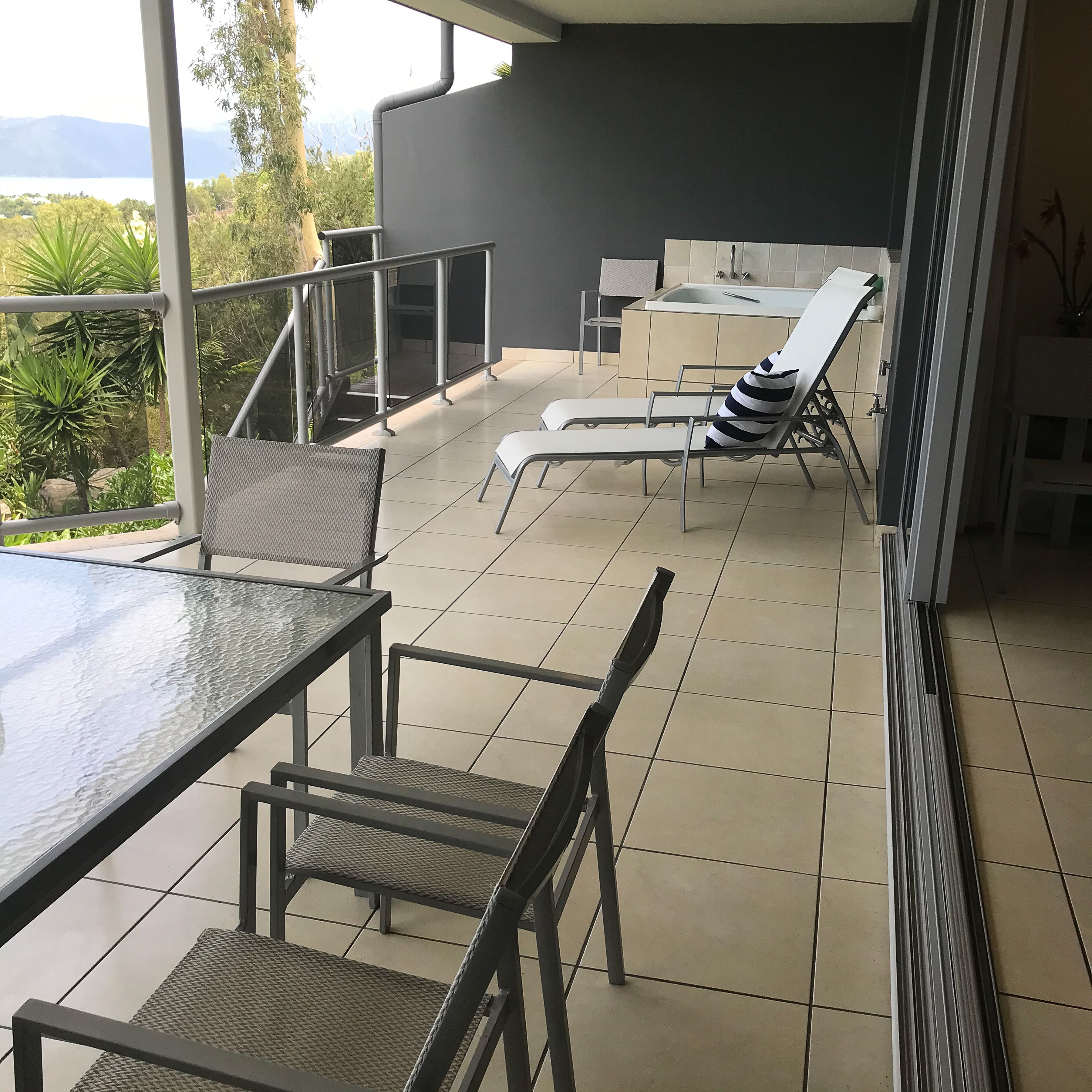 Patio esterno moderno con tavolo in vetro, sedie in metallo e una sdraio.