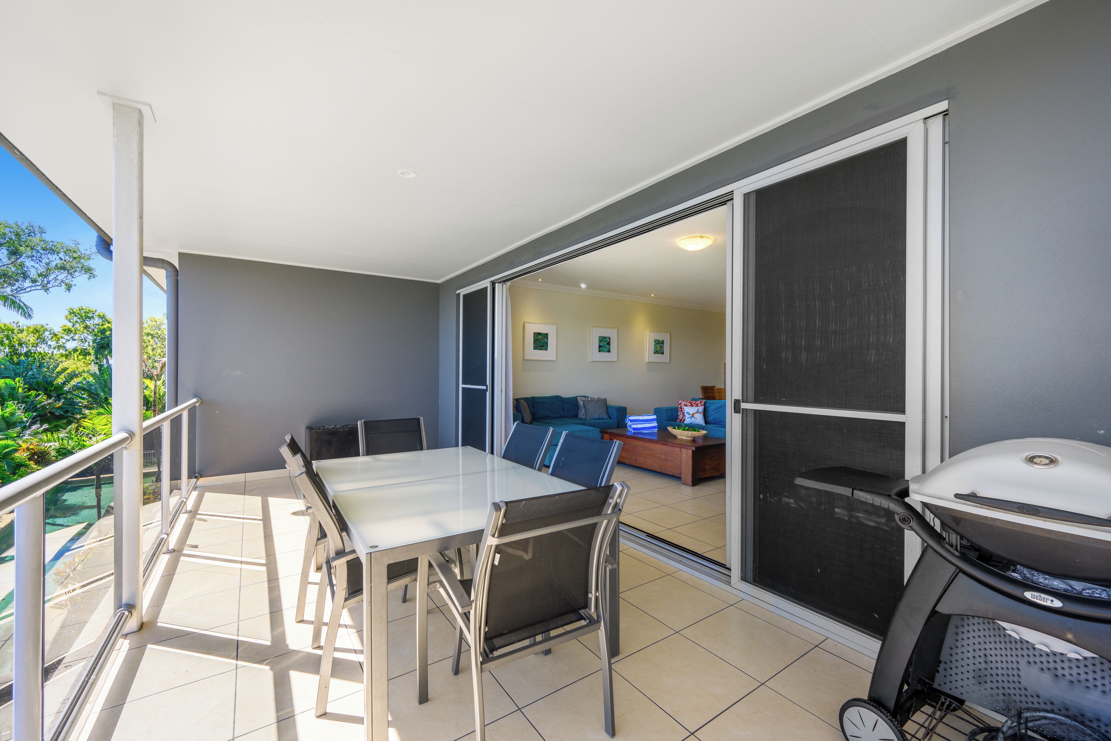 A balcony with a dining table and chairs, a barbecue grill, and a sliding glass door leading to a living area with a sofa and coffee table.