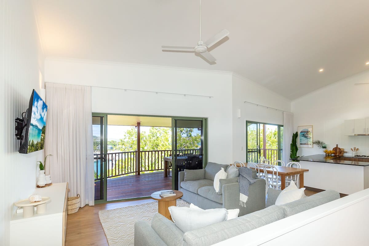 A modern living room with a sofa, coffee table, and a dining area with a table and chairs. There is a balcony with a view of the outdoors.