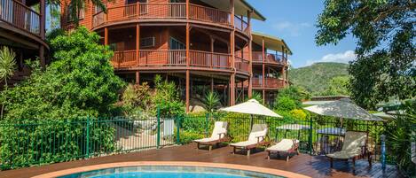 A pool area with lounge chairs and umbrellas, surrounded by a wooden deck and a fenced garden.