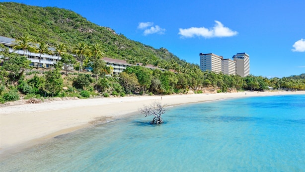 A beach with clear turquoise water, a sandy shore, and a building with multiple floors in the background. - Heliconia Grove 9 (Whitsundays)