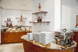 A buffet setup with stacked plates, a coffee station, and shelves with bottles and glasses.