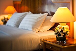 A neatly made bed with white linens and pillows, a bedside table with a lamp and a vase of flowers, and a wooden headboard.
