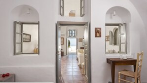 A hallway with a bed, a wooden chair, a small table with red objects, and a bathroom with a mirror and sink. - Nautilus (Santorini)