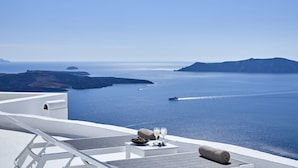 A seaside terrace with a white lounge chair, a table with a bowl of fruit and two glasses, and a rolled towel. - Nautilus (Santorini)