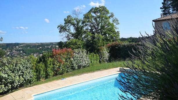 A swimming pool with a clear blue water, surrounded by a garden with various plants and a view of a distant town. - Impressive villa overlooking the hill (Joyeuse)