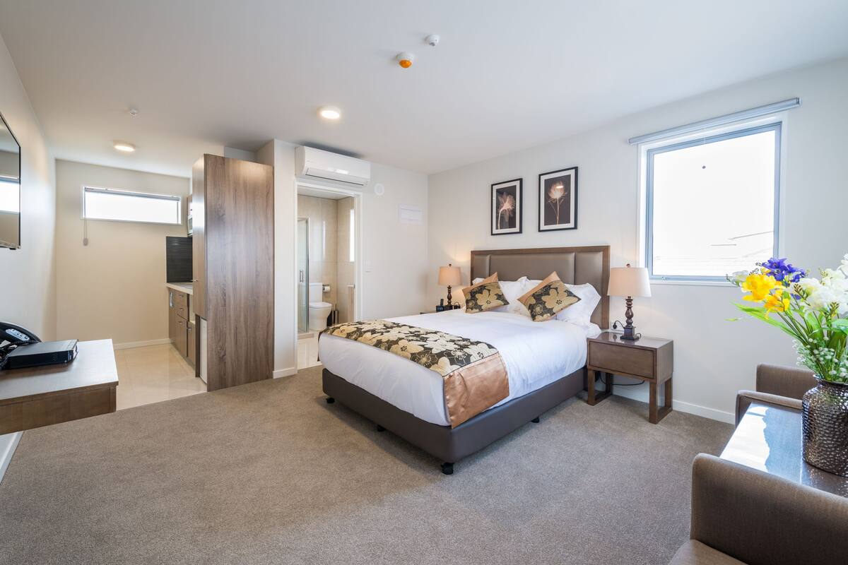 A hotel room with a large bed, bedside tables, a desk, a chair, a TV, and a window with flowers.
