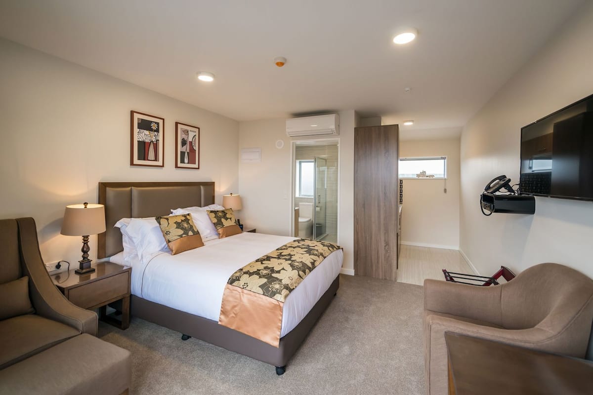 A hotel room with a bed, bedside tables, a desk, a chair, a TV, and a bathroom visible through an open door.