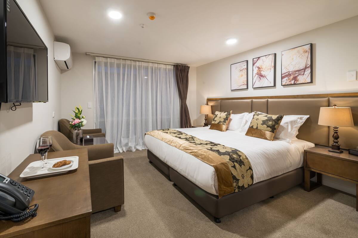 A hotel room with a large bed, a desk with a phone and a plate of food, a chair, and a TV mounted on the wall.