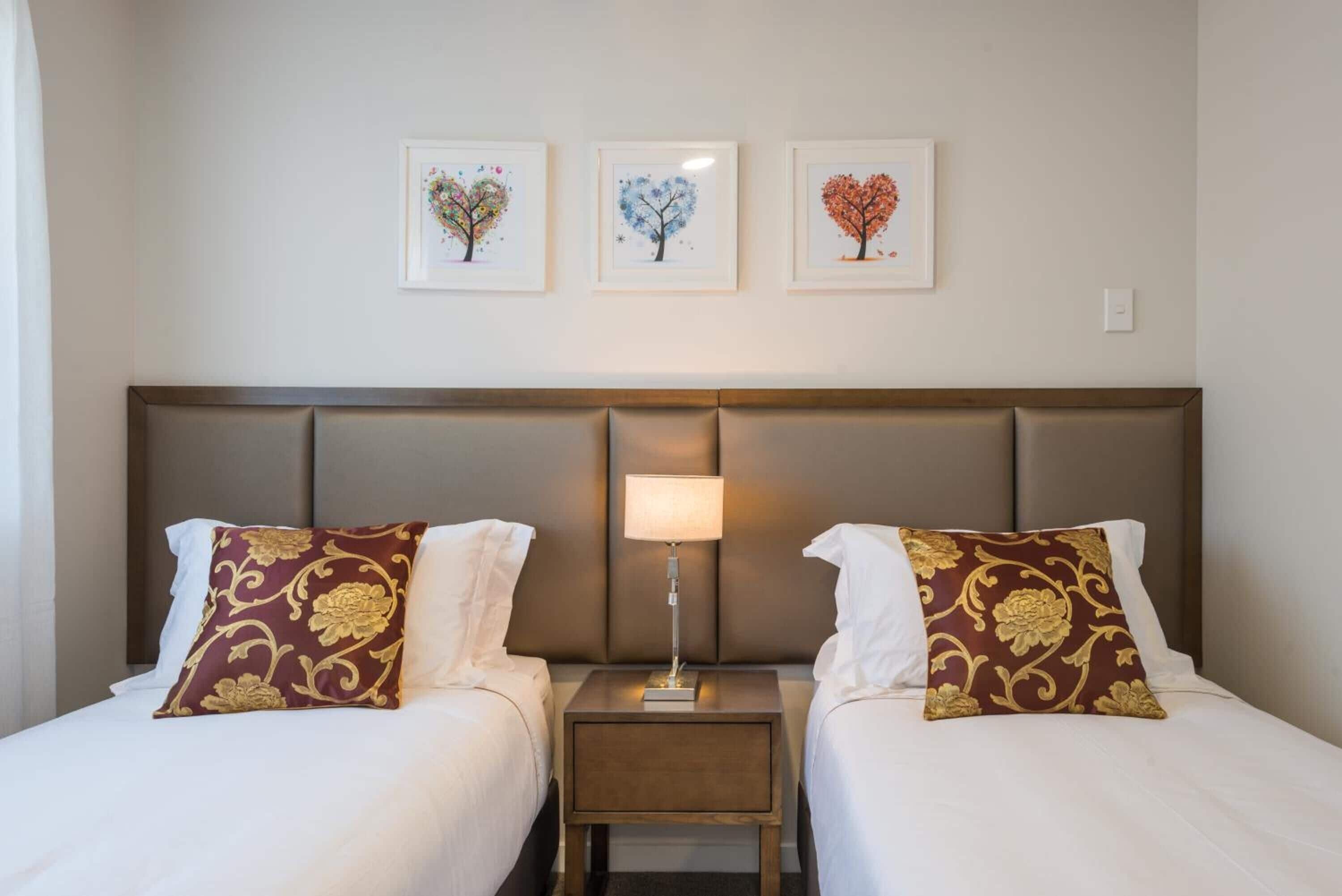 A hotel room with two beds, a nightstand with a lamp, and three framed pictures on the wall.