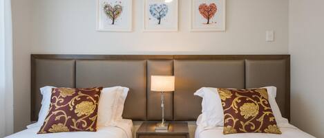 A hotel room with two beds, a nightstand with a lamp, and three framed pictures on the wall.