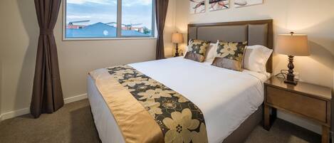 A neatly made bed with a floral bedspread, a bedside lamp, and a view of the outside through a window with curtains.