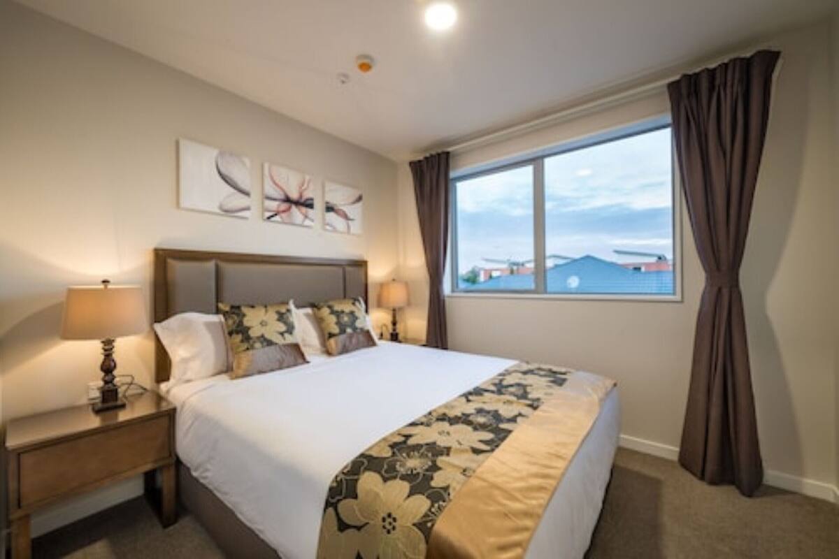 A hotel room with a large bed, a nightstand with a lamp, a window with curtains, and artwork on the wall.