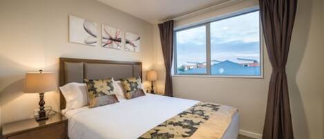 A hotel room with a large bed, a nightstand with a lamp, a window with curtains, and artwork on the wall.
