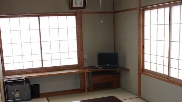 Una habitación japonesa tradicional con suelo de tatami, una mesa de madera baja, un televisor sobre un mueble y un marco de fotos en la pared.