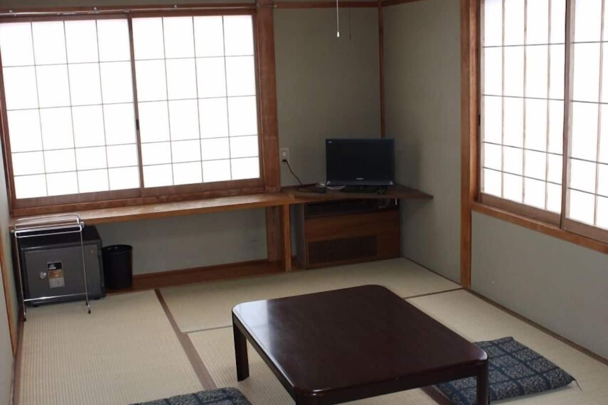 A traditional Japanese room with tatami flooring, a low wooden table, a television on a stand, and a picture frame on the wall.