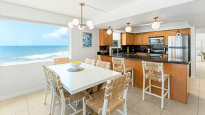 A modern kitchen with a view of the ocean, a dining area with wicker chairs, and a white table. - Paradise Beach Club - Oceanfront and Penthouse Deluxe Condos with Heated Pool (Satellite Beach)